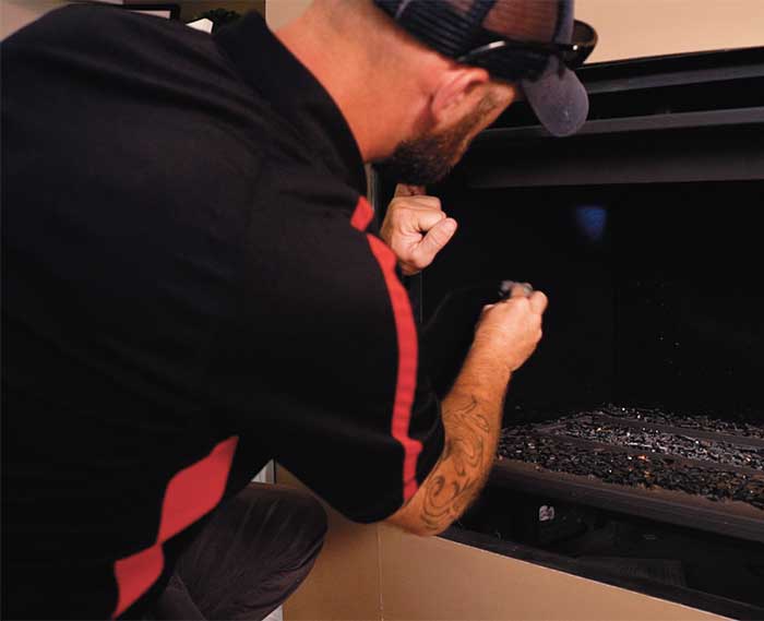 man with black baseball cap and shirt with red trim using a flashlight to inspect a gas fireplace